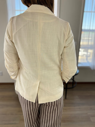 A person viewed from the back wearing a light cream linen-blend blazer with a center back seam and striped brown pants, standing in a brightly lit room with wood floors and windows.