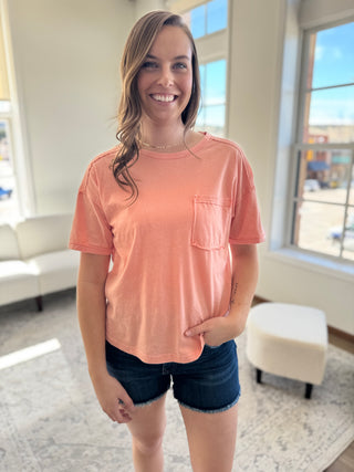 Lady standing wearing peach washed tee with front pocket and denim shorts.