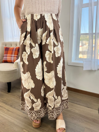 A person wearing loose, wide-leg brown pants with a large cream floral print and a decorative border along the hem, paired with a cream knit top and beige slide sandals in a bright indoor room.