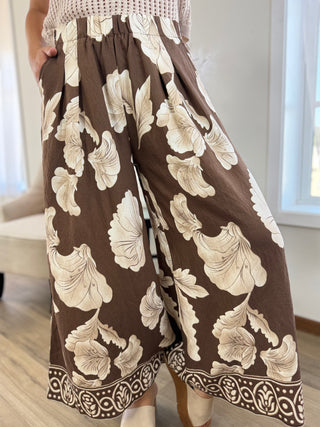 A person wearing wide-leg cropped pants in brown with a large beige floral print and a decorative border along the hem, shown indoors near a window.