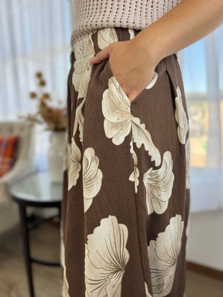 A close-up of a person wearing brown wide-leg pants with a large cream floral print, standing in a bright indoor setting.