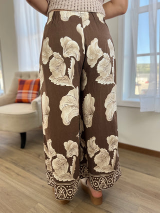 A person wearing brown wide-leg pants with a large cream floral print and a decorative patterned hem, standing in a bright room with wood floors and a window.
