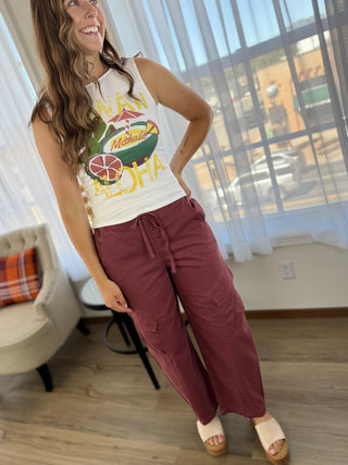 A woman modeling a sleeveless white tank top with a colorful Hawaiian graphic and matching dusty rose wide-leg drawstring pants, standing in a bright room with large windows and sheer curtains.