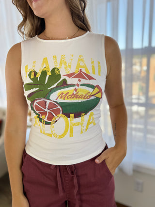 A woman wearing a white sleeveless tank top with a colorful distressed graphic that reads 'HAWAII ALOHA' in yellow, featuring green palm leaves, a coconut shell, and a red-and-yellow beach umbrella, paired with burgundy drawstring shorts in a bright indoor setting.