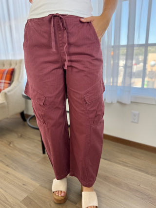 A person wearing loose, wide-leg cargo-style pants in a dusty rose or mauve color, paired with a white top and white slide sandals, standing indoors on a wood floor near a window.