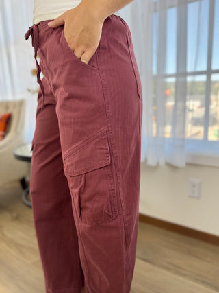 A person wearing dusty rose cargo pants with side pockets, standing in a brightly lit room with a window and light wood floor.