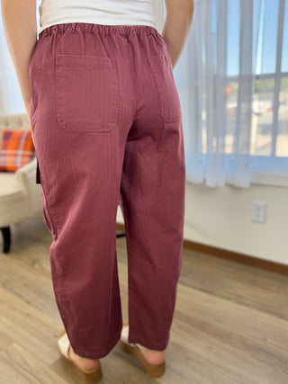 A person shown from the back wearing loose, wide-leg pants in a dusty rose or mauve color, paired with a white top and tan slide sandals in a home interior setting.