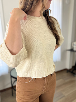 A woman wearing a cream-colored ribbed knit short-sleeve sweater and brown corduroy pants, standing in a brightly lit room.