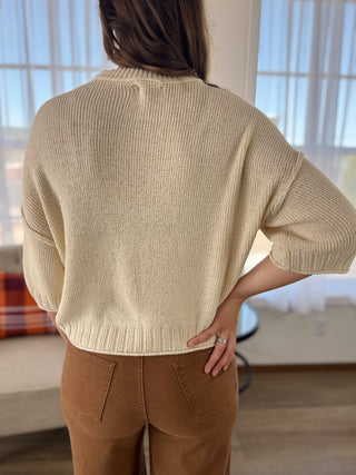 A person shown from the back wearing a cream-colored ribbed knit sweater and brown pants in a bright indoor setting.
