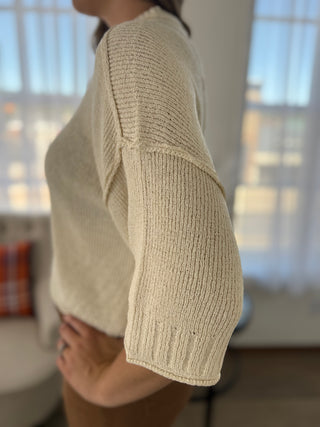 A close-up side view of a person wearing a cream-colored ribbed knit sweater in a bright, modern room with soft window light.