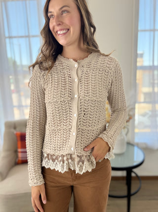 A woman wearing a cream-colored, textured crochet cardigan with a lace hem and button-front closure, paired with brown pants, standing in a brightly lit room.