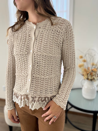 A woman wearing a beige chunky knit cardigan with a lace hem, paired with brown pants, standing in a bright indoor setting.