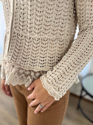 A close-up of a person wearing a beige open-knit long-sleeve cardigan with a ruffled hem over a matching lace top, paired with brown pants.