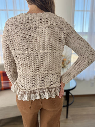 A woman viewed from behind wearing a beige chunky knit sweater with a ruffled hem, paired with brown pants, in a brightly lit indoor room.