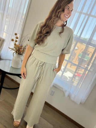 A woman modeling a light sage green two-piece lounge set with a textured waffle pattern, featuring a short-sleeve top and matching wide-leg pants with a drawstring waist, in a bright room with sheer curtains.