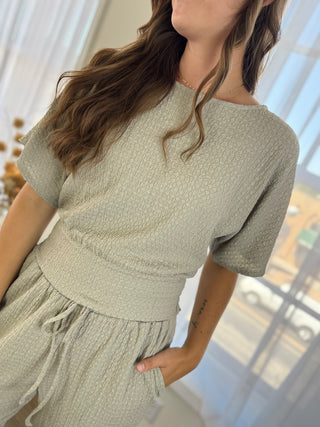 A woman wearing a light sage green textured knit two-piece set consisting of a short-sleeve top and matching drawstring shorts, photographed in a bright indoor setting.