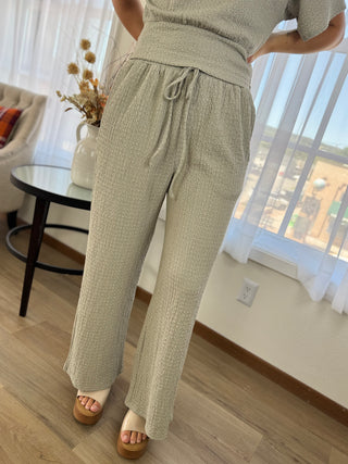 A person wearing a light beige textured two-piece outfit with matching drawstring pants and a short-sleeve top, standing in a bright room with wood floors, sheer curtains, and a small round table.