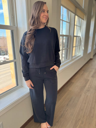 A person standing by a window wearing black lounge pants with a thick elastic waistband and drawstring, with side pockets and wide leg.