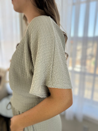 A woman wearing a textured sage green short-sleeve top, shown from a side-back angle in a bright room with sheer curtains.