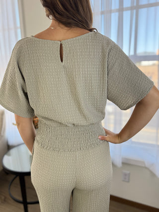 A woman shown from the back wearing a textured two-piece jumpsuit in a light sage green color, featuring a smocked waist and short flutter sleeves.