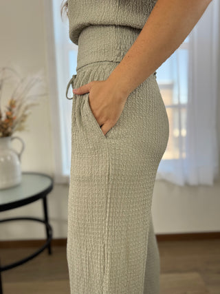 A close-up of a person wearing a textured, light sage green wide-leg skirt or pants with side pockets, standing in a bright room.