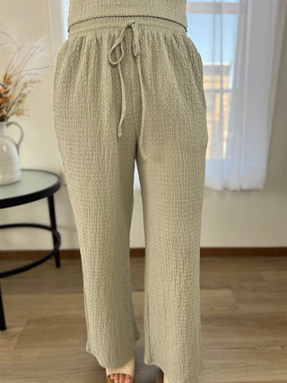 A person wearing textured wide-leg drawstring pants in a light beige or oatmeal color, standing indoors on a wood floor near a window.