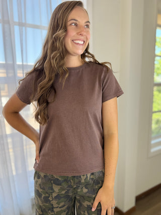 A woman smiling and looking to the side, wearing a short-sleeve crewneck t-shirt in a washed brown color, paired with green and brown camouflage pants, standing in a brightly lit room.