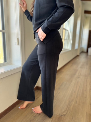 A person standing by a window wearing black lounge pants with a thick elastic waistband and drawstring, with side pockets and wide leg.