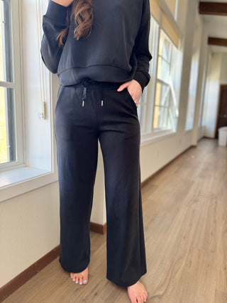 A person standing by a window wearing black lounge pants with a thick elastic waistband and drawstring, with side pockets and wide leg.
