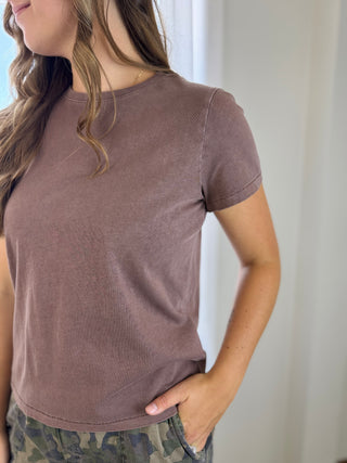 A woman wearing a short-sleeve crewneck t-shirt in a washed, muted mauve-brown color, paired with camouflage pants, shown from the side against a bright indoor background.