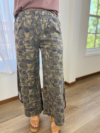 A person wearing loose camouflage wide-leg pants with an elastic waistband, shown from the waist down in a bright indoor room with wood flooring and a window.