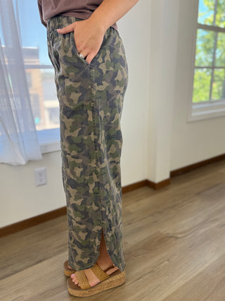 A person wearing long camouflage pants with a side slit and side pocket, standing indoors on a wood floor near windows.