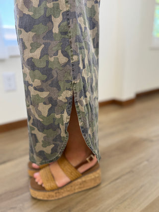 Close-up of a person's lower legs wearing camouflage print wide-leg jeans with a front slit and tan platform wedge sandals on a wooden floor.