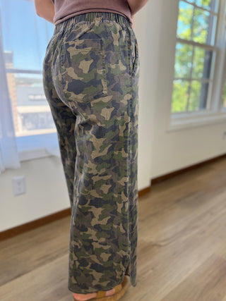 A person wearing camouflage wide-leg pants, shown from the back in an indoor room with windows and wood flooring.