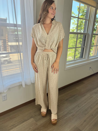 A woman modeling a matching beige two-piece set with tiny black polka dots, featuring a short-sleeve wrap-style top and wide-leg pants, standing in a room with large windows and wood floors.
