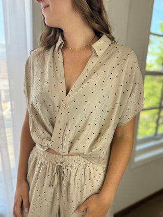 A woman wearing a beige short-sleeve button-front top and matching drawstring pants set, both with small black polka dots, photographed indoors near a window.