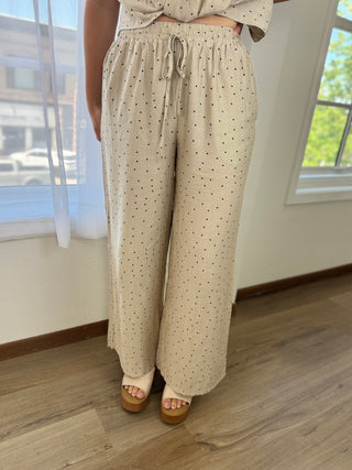 A person wearing loose, wide-leg drawstring pants in a light beige or cream tone with small evenly spaced black polka dots, standing indoors on a wood floor near windows.