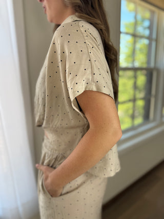 A woman shown from the side in a home setting, wearing a beige short-sleeve top and matching pants set with a small black polka dot pattern.