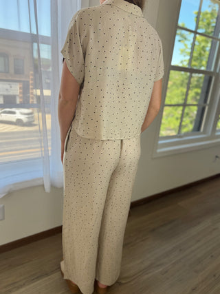 A person shown from the back wearing a matching short-sleeve button-up shirt and wide-leg pants set in a light beige fabric with small black polka dots, standing in a room with large windows and wood flooring.