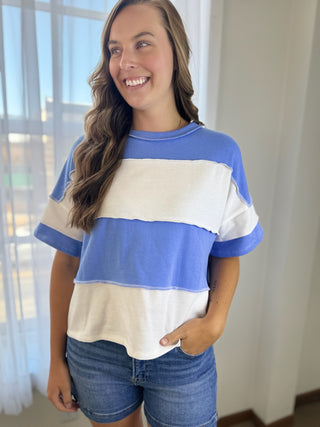 A woman wearing a short-sleeve color-blocked t-shirt with horizontal stripes of royal blue and off-white, paired with blue denim shorts, standing in a brightly lit room.
