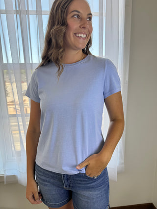 A woman wearing a light periwinkle blue short-sleeve crewneck t-shirt and blue denim shorts, standing in front of a window with sheer white curtains.