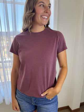 A woman wearing a short-sleeve crewneck t-shirt in a dusty mauve or plum color, paired with blue jeans, standing indoors near a window.