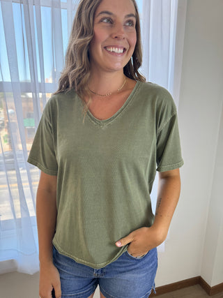 A woman wearing a short-sleeve, v-neck olive green t-shirt and blue denim shorts, standing in a brightly lit room with sheer white curtains.