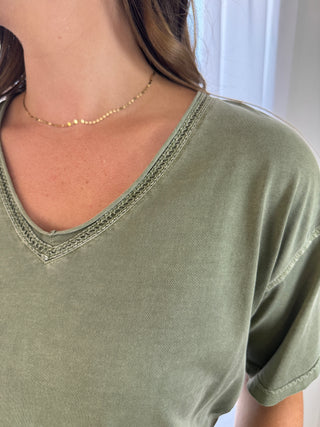 Close-up of a person wearing an olive green short-sleeve V-neck T-shirt with a delicate gold chain necklace, shown against a soft light background.