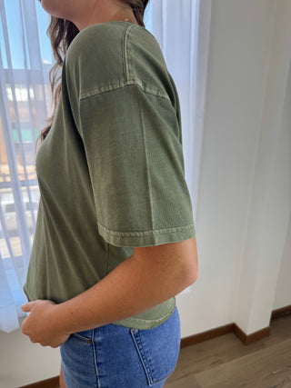 A woman wearing a cropped, short-sleeve olive green t-shirt and blue denim shorts, standing in a brightly lit room with white walls and a window.