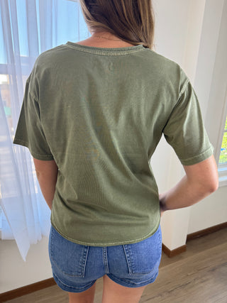 A woman viewed from behind wearing an olive green short-sleeve ribbed t-shirt and blue denim shorts in a brightly lit room.