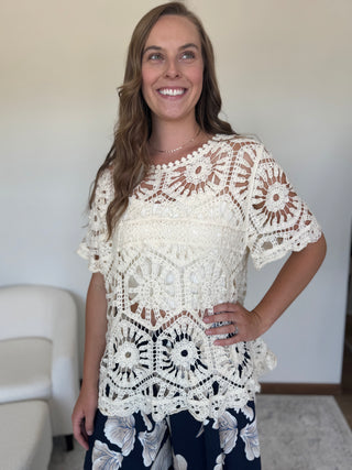 A smiling woman wearing a cream-colored, short-sleeved crochet lace top and navy blue floral pants, standing in a brightly lit indoor room.