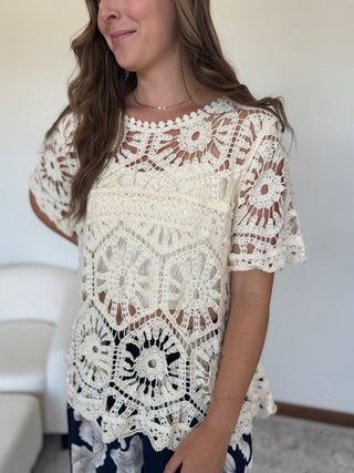 A woman wearing a cream-colored, short-sleeved crochet lace top with intricate floral patterns, paired with dark patterned pants, standing in a brightly lit room.