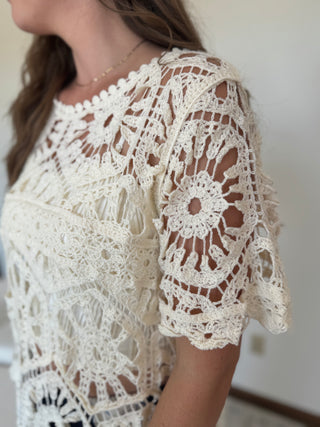 A close-up of a woman wearing a cream-colored, hand-crocheted lace top with short flutter sleeves and an intricate floral pattern.