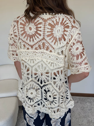A woman viewed from behind wearing a cream-colored, short-sleeved crochet lace top over dark navy and white patterned pants, standing indoors.